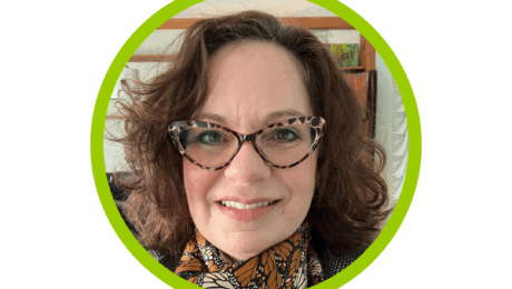 headshot of Alexia Georghiou, smiling, wearing cat eye style glasses with a monarch butterfly scarf and black and white checkered blazer